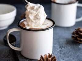 Side view of whipped cream being spooned onto a mug of hot chocolate.