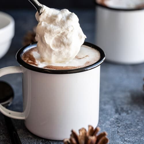 Side view of whipped cream being spooned onto a mug of hot chocolate.