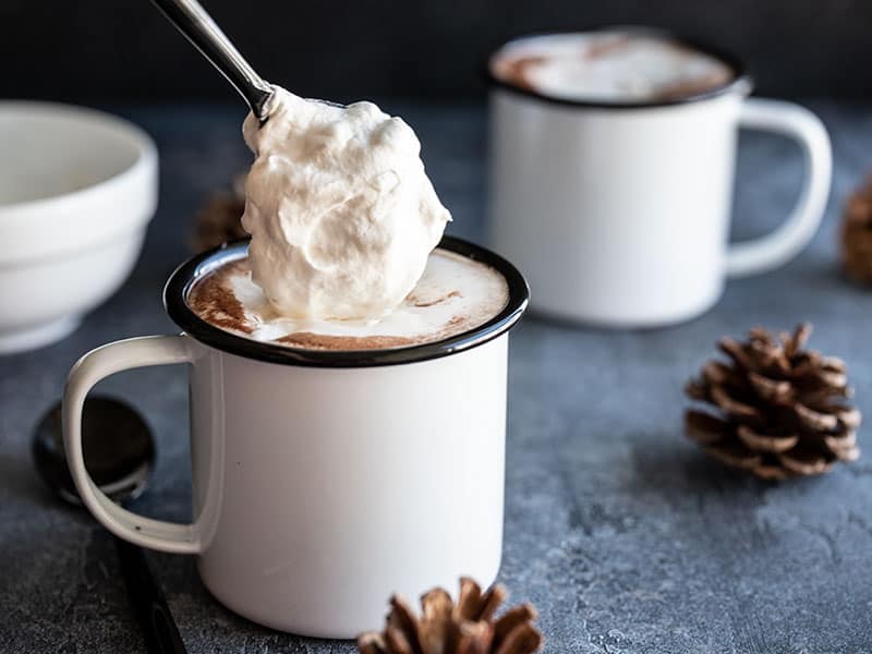 Homemade whipped cream being spooned onto a mug of hot cocoa, a second mug in the background