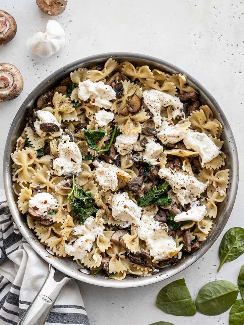 A skillet full of Mushroom and Spinach Pasta with Ricotta with spinach leaves and whole mushrooms on the sides