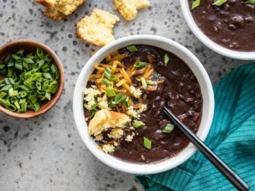 Two bowls of Smoky Black Bean Soup with various toppings, next to a bowl of sliced green onion and a crumbled corn muffin.