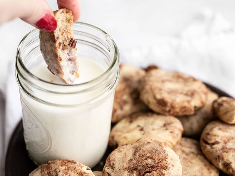 A cinnamon pecan sandie being dunked into a glass of milk on a plate full of cookies