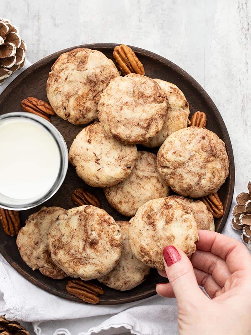 Cinnamon pecan sandies on a wooden plate with a glass of milk, a hand picking up one cookie