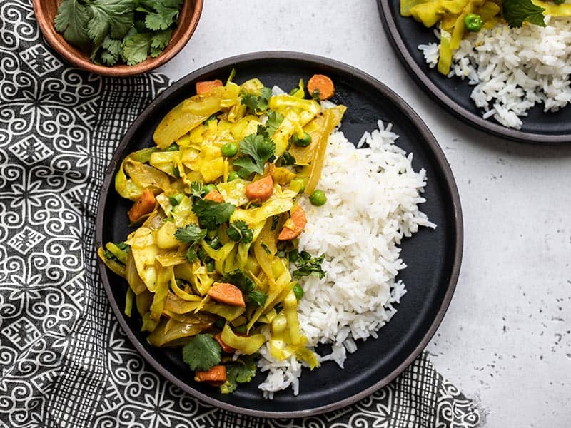 A black plate with rice and curried cabbage with a bowl of cilantro on the side