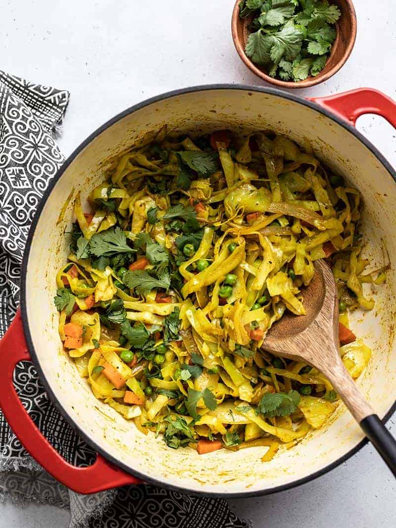 Curried cabbage in a red pot with a wooden spoon and garnished with fresh cilantro