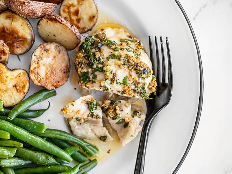 A piece of Garlic Butter Baked Chicken on a plate with potatoes, green beans, and a black fork.