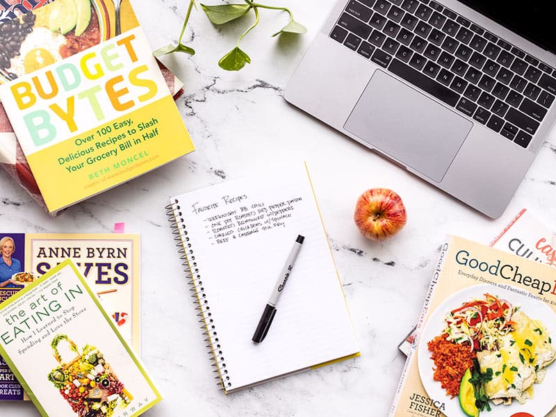 A notebook with a list of recipes surrounded by cookbooks and a laptop.