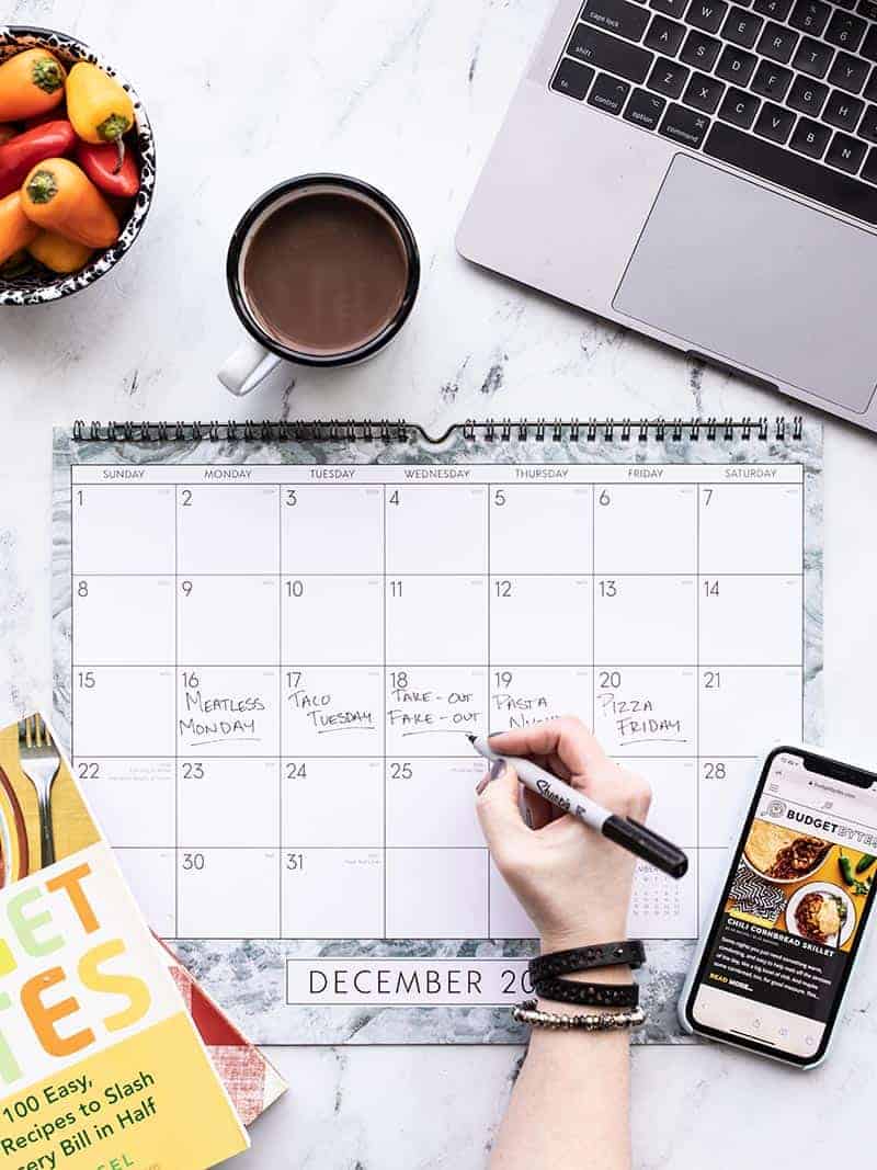 Hand writing on a large calendar with cookbooks, cell phone, and laptop on the sides