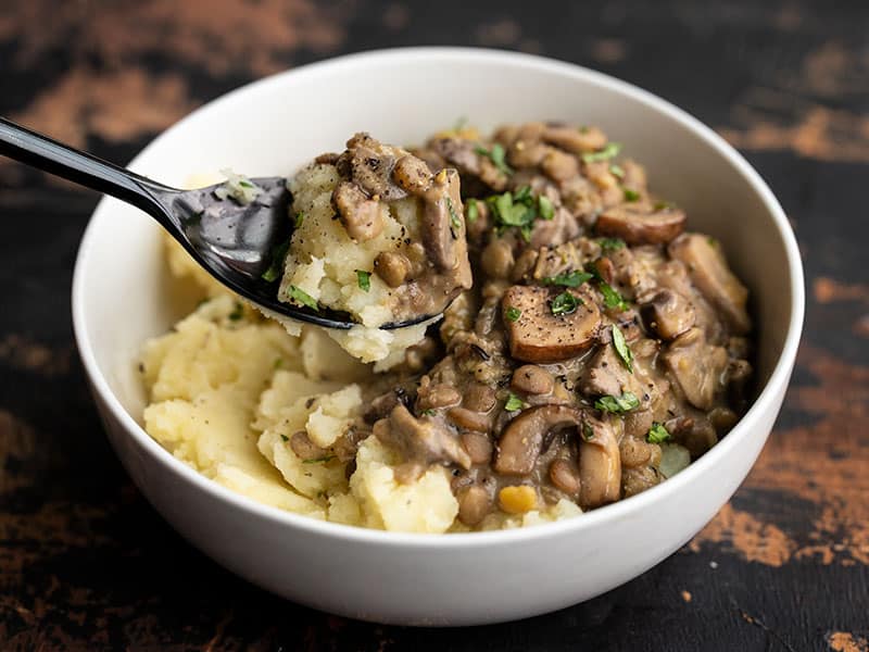 A bowl of Lentils with Creamy Mushroom Gravy over mashed potatoes, a bite being lifted by a black spoon.