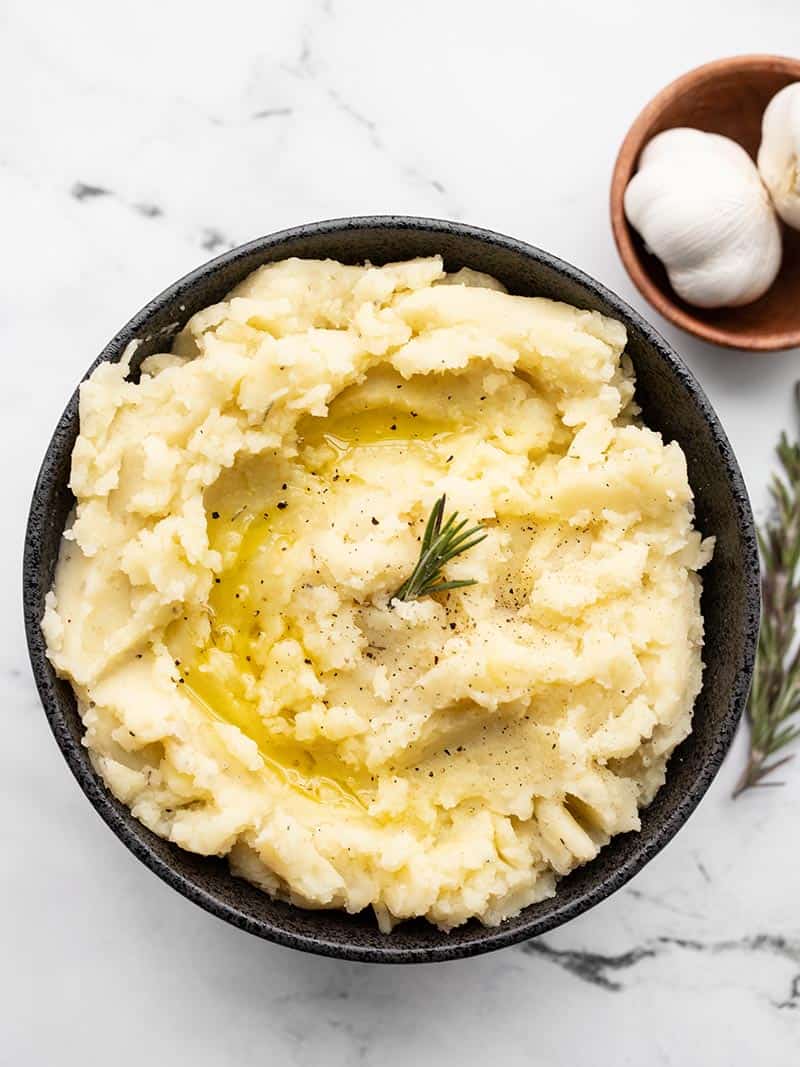 Olive Oil Mashed Potatoes in a black bowl garnished with fresh rosemary and garlic bulbs on the side