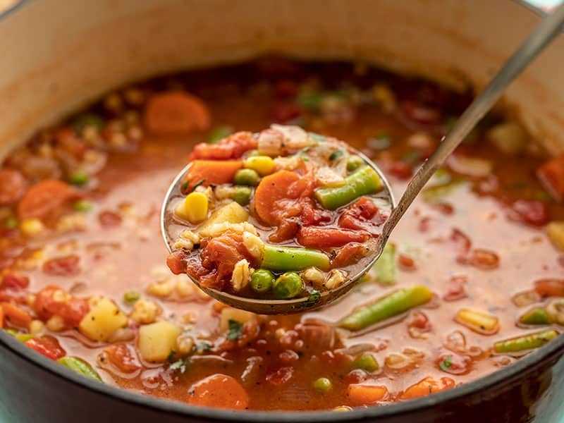 Front view of a ladle full of Vegetable Barley Soup hovering over the pot full of soup.