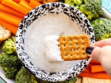 A cracker being dipped into the whipped cottage cheese dip, the platter of vegetables and crackers in the background