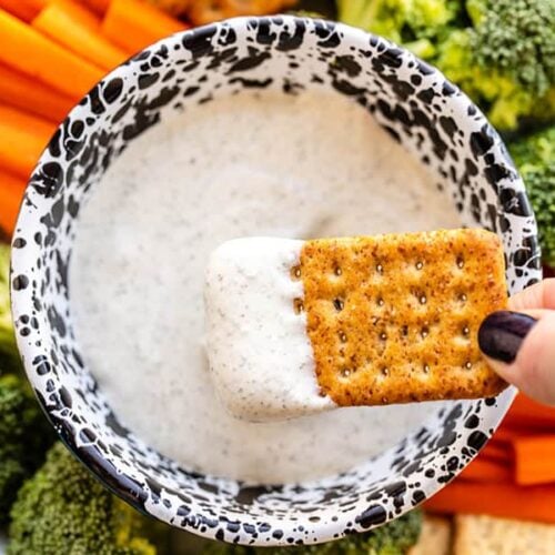A cracker being dipped into the whipped cottage cheese dip, the platter of vegetables and crackers in the background