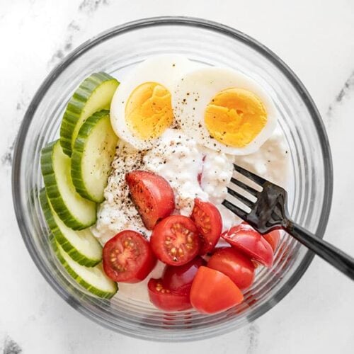 A glass bowl with cottage cheese, hard boiled egg, tomatoes, cucumber, and pepper.