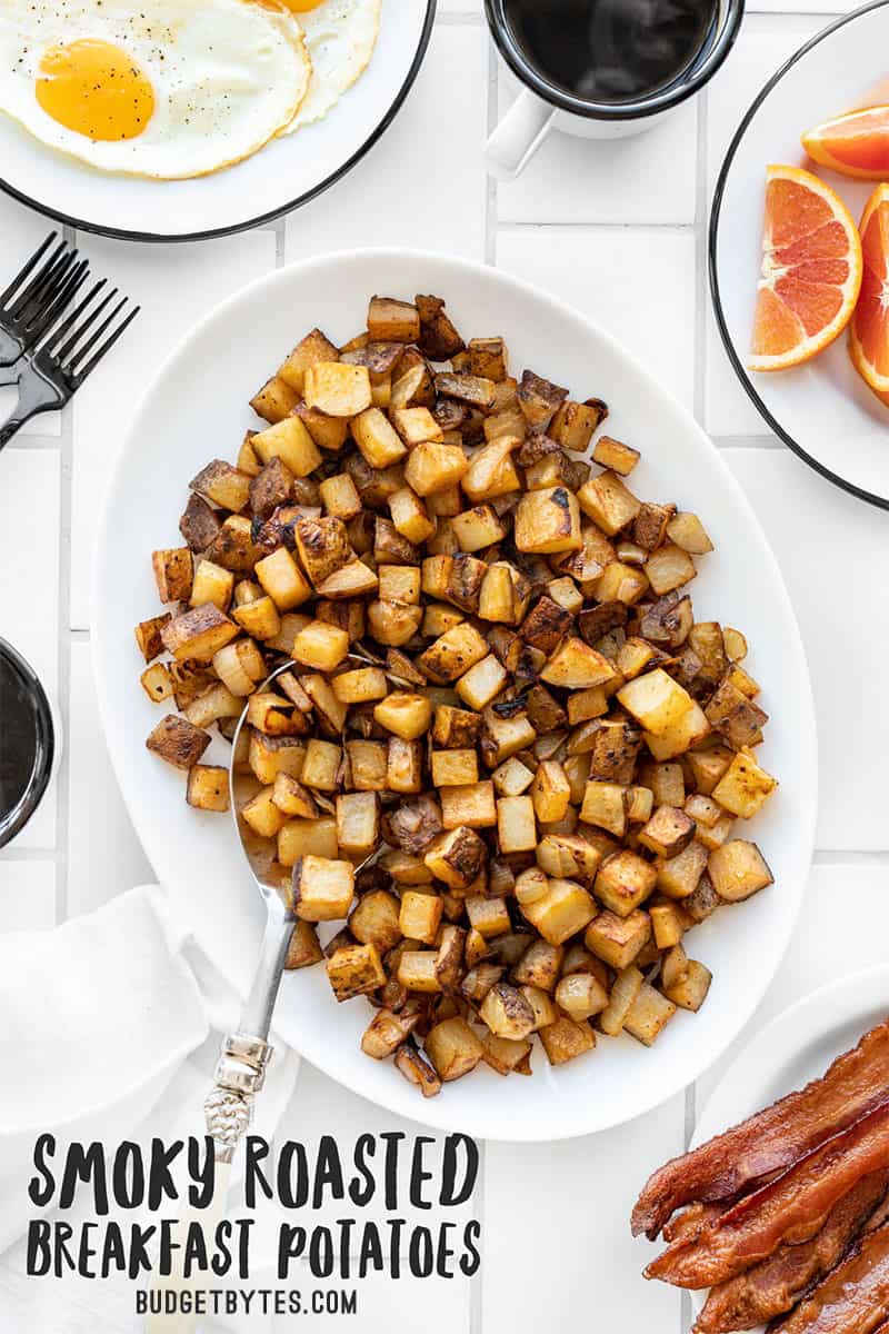 A platter full of breakfast potatoes with coffee, eggs, bacon, and orange slices on the side