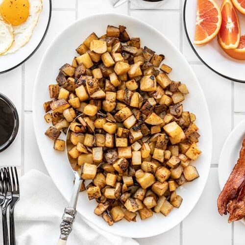 A platter full of roasted breakfast potatoes with eggs, coffee, oranges, and bacon on the side