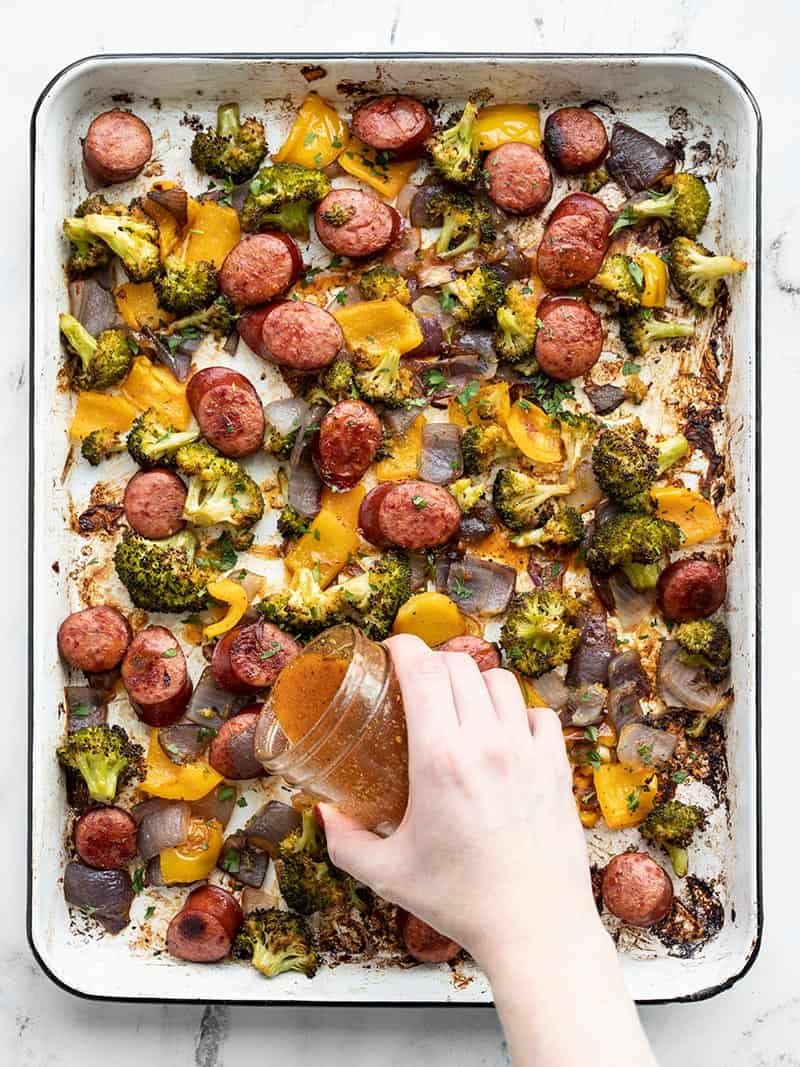 A hand drizzling smoky vinaigrette over a sheet pan full of roasted sausage and vegetables