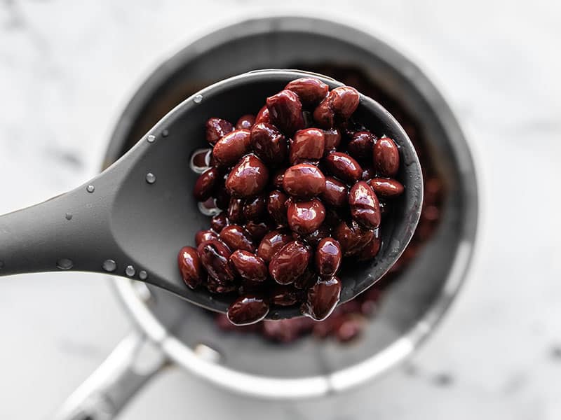 Warmed black beans in a sauce pot, a spoon lifting some toward the camera