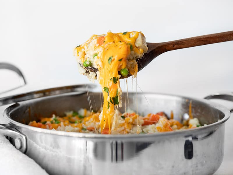 A wooden spoon lifting chicken and rice out of the skillet with melted cheese stretching between