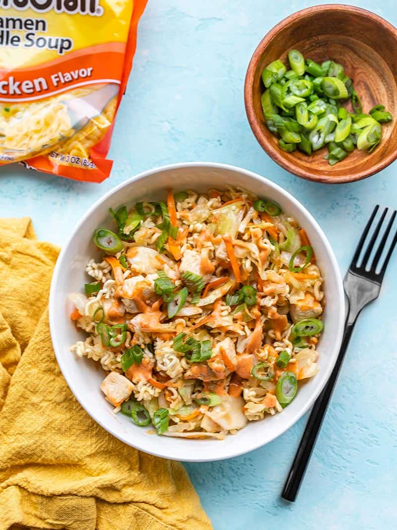 A bowl of crunchy chicken ramen stir fry next to a ramen package, yellow napkin, and a bowl of sliced green onions