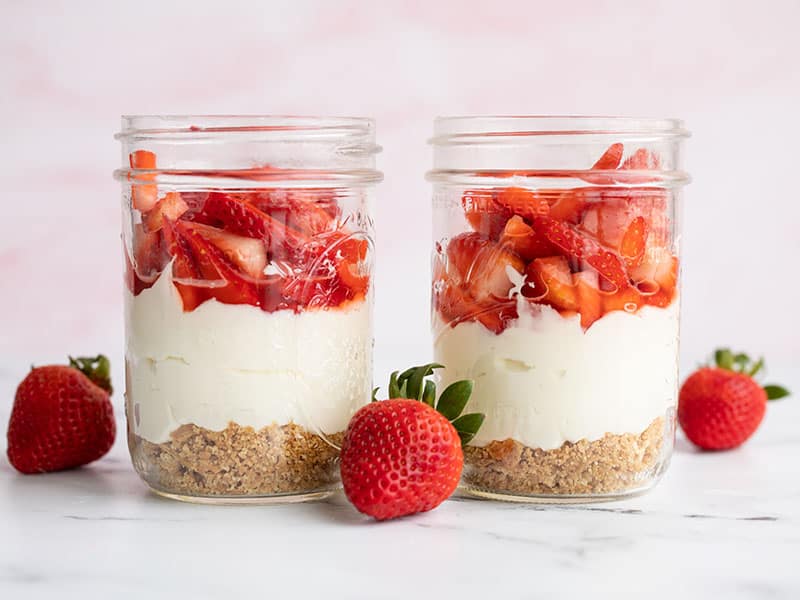 Two glasses of unbaked strawberry cheesecake side by side with whole strawberries on the side