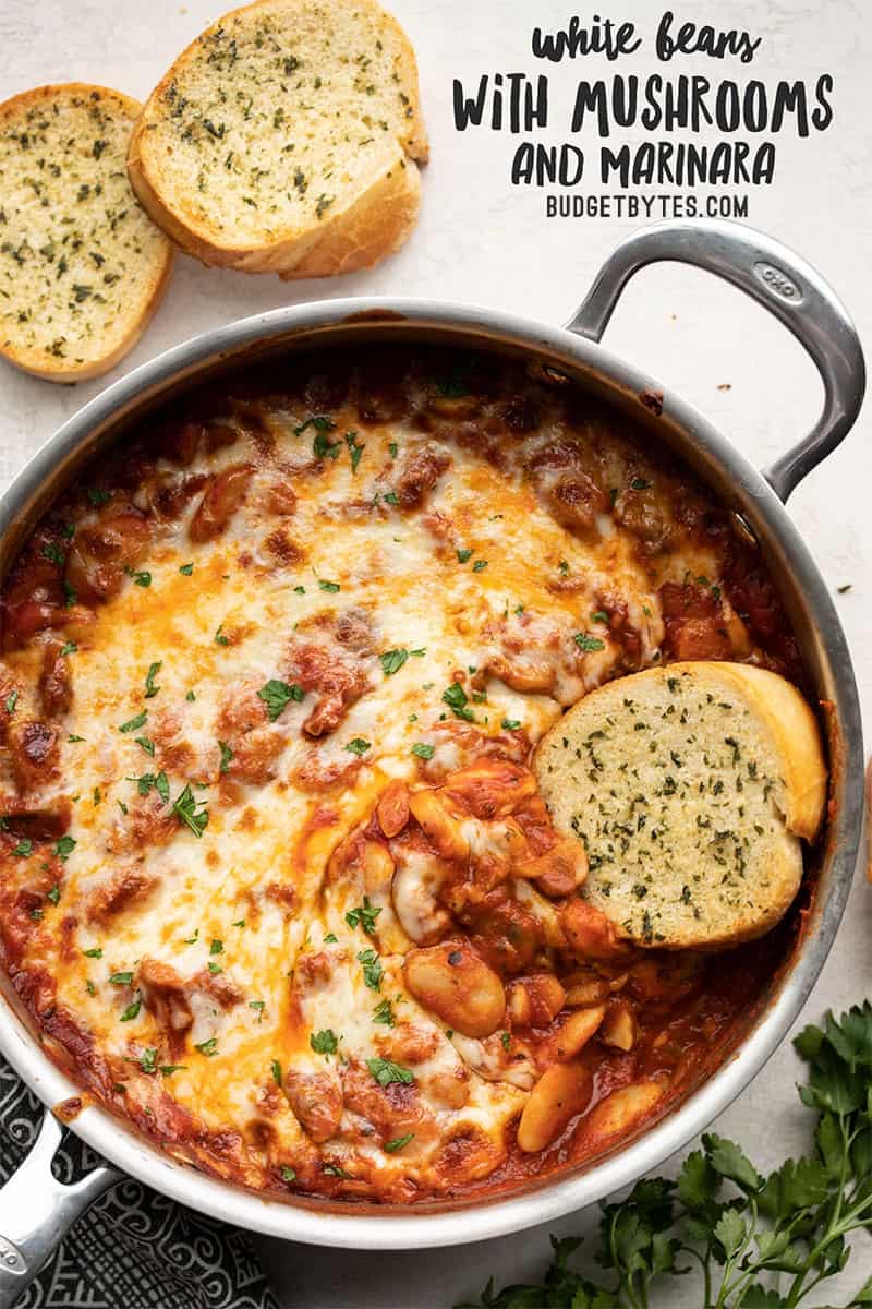 Overhead view of a skillet full of white beans with mushrooms and marinara, title text at the top