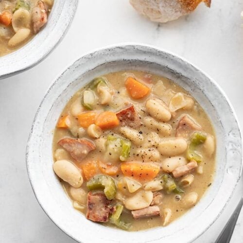 Two bowls of chunky ham and bean soup with chunks of bread on the side