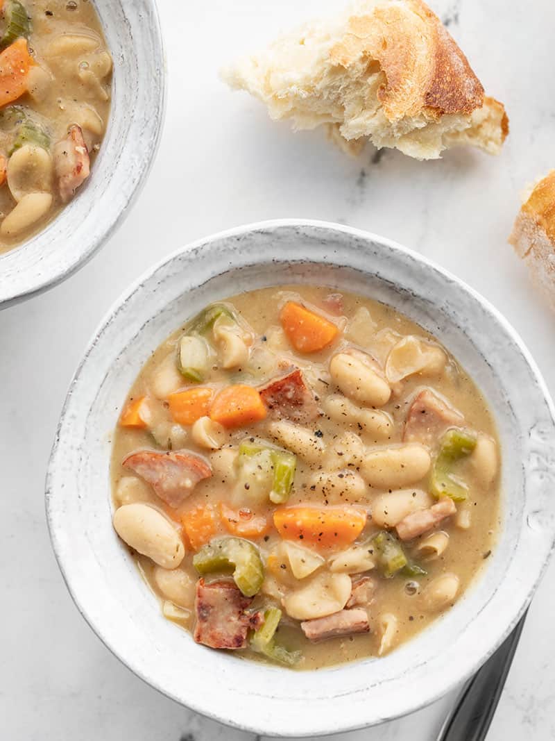 Two bowls of chunky ham and bean soup with torn chunks of bread on the side