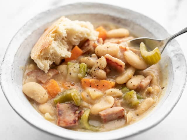 Close up front view of a bowl full of chunky ham and bean soup with a spoon lifting some and a chunk of bread in the side of the bowl.