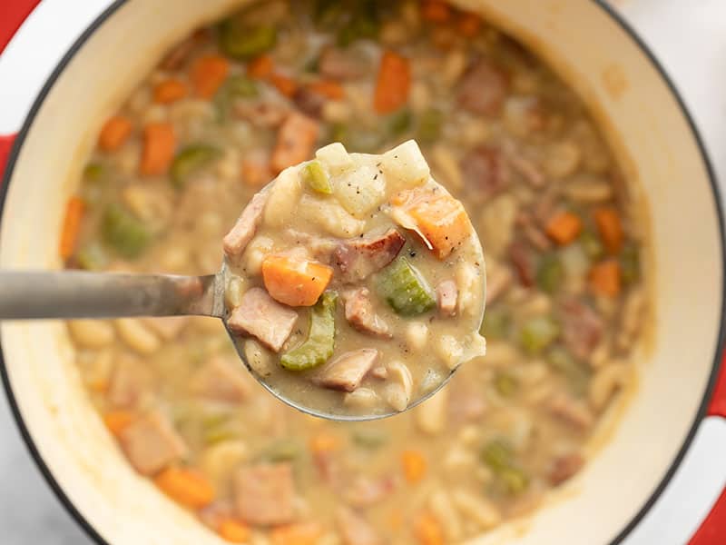 Close up of a ladle full of chunky ham and bean soup being held over the soup pot