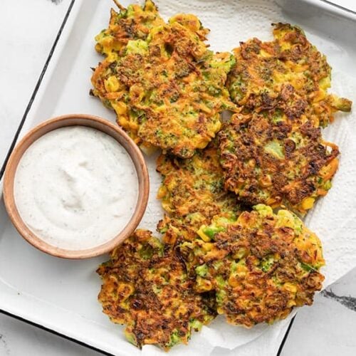 Overhead view of a white tray lined with paper towel, with vegetable fritters and a small bowl of garlic herb dipping sauce