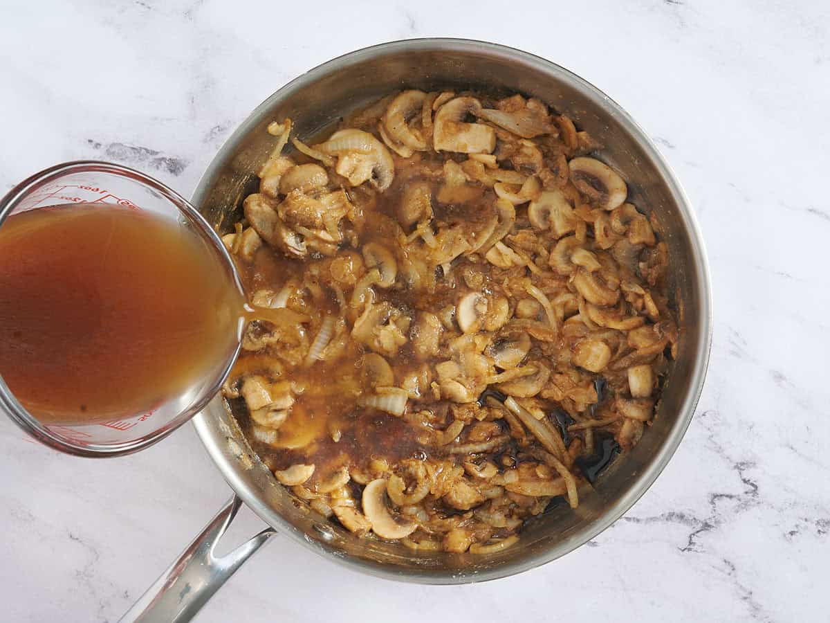 Broth being poured into a skillet with sliced onions and mushrooms.