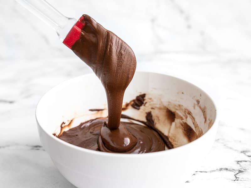 Finished chocolate icing dripping off a red spatula into the bowl