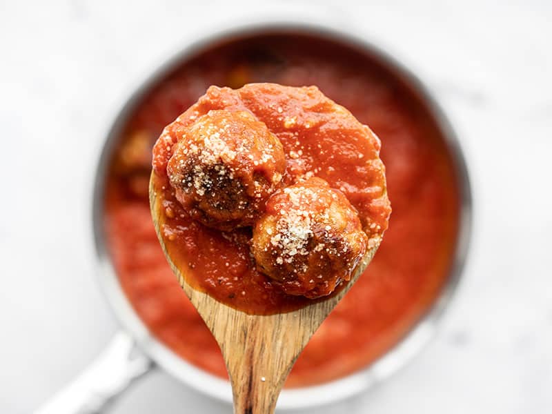 Two chicken Parmesan meatballs on a wooden spoon held above the pot of sauce.