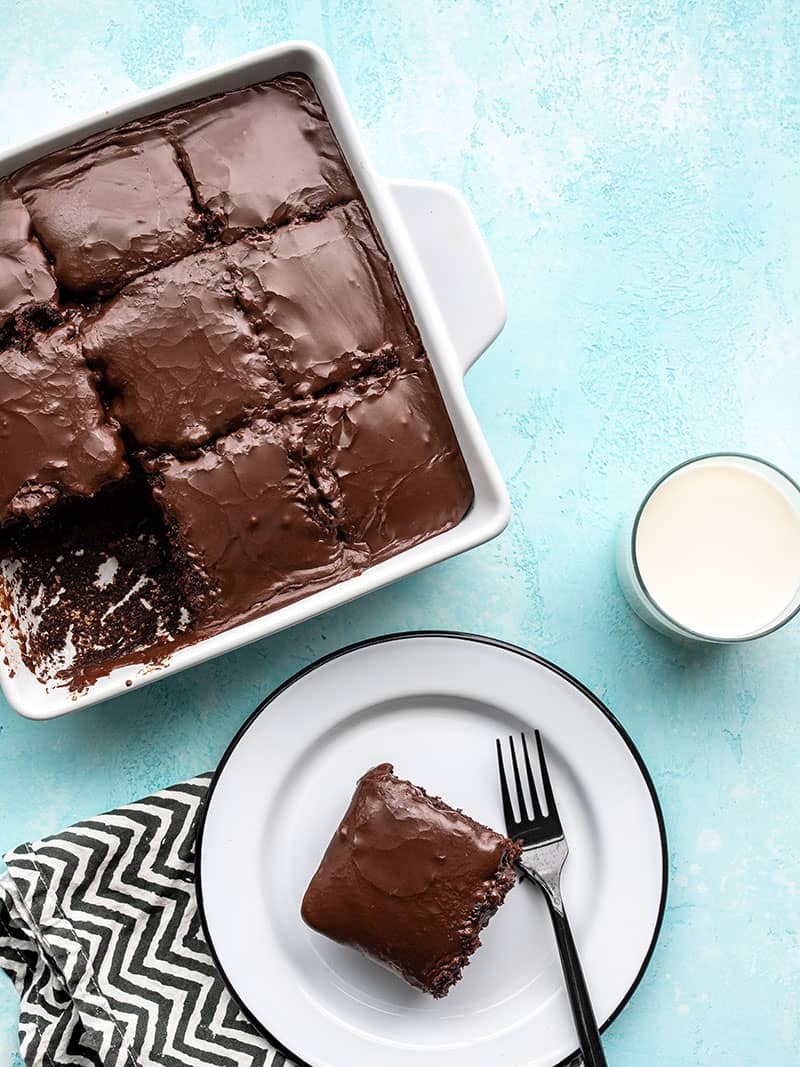 A white casserole dish with chocolate cake, one slice on a white plate and a glass of milk near by.