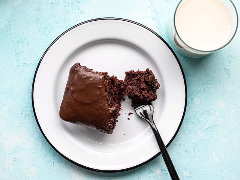 A piece of chocolate depression cake on a white plate with a fork and a glass of milk on the side