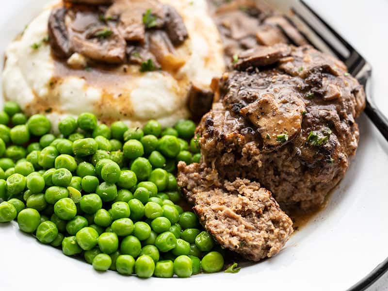 Close up side view of a Salisbury Steak on a plate with peas and mashed potatoes, and a piece of the steak cut off the side.