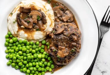 Um prato cheio de Salisbury Steak com molho de cogumelos, purê de batatas e ervilhas.