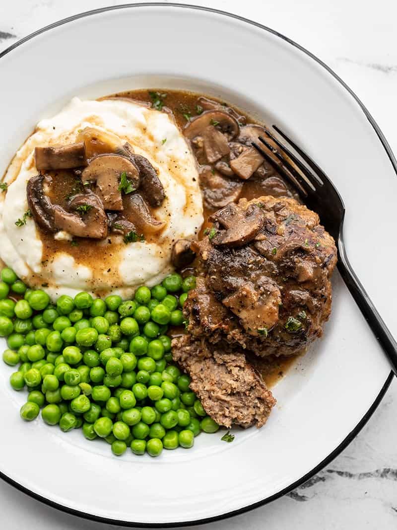 Salisbury steak with mushroom gravy on a plate with mashed potatoes and peas, a fork on the side.