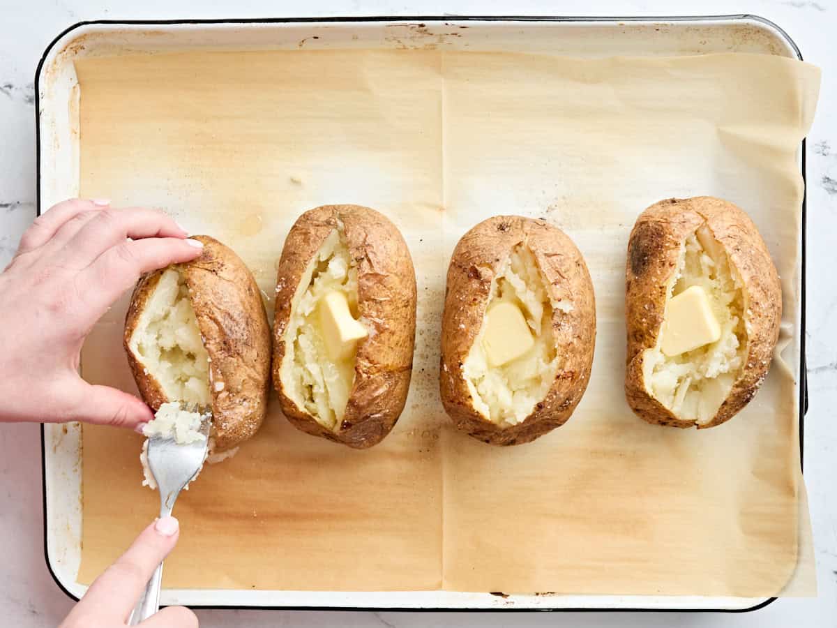 A fork fluffing up the inside of a baked potato on a baking sheet.