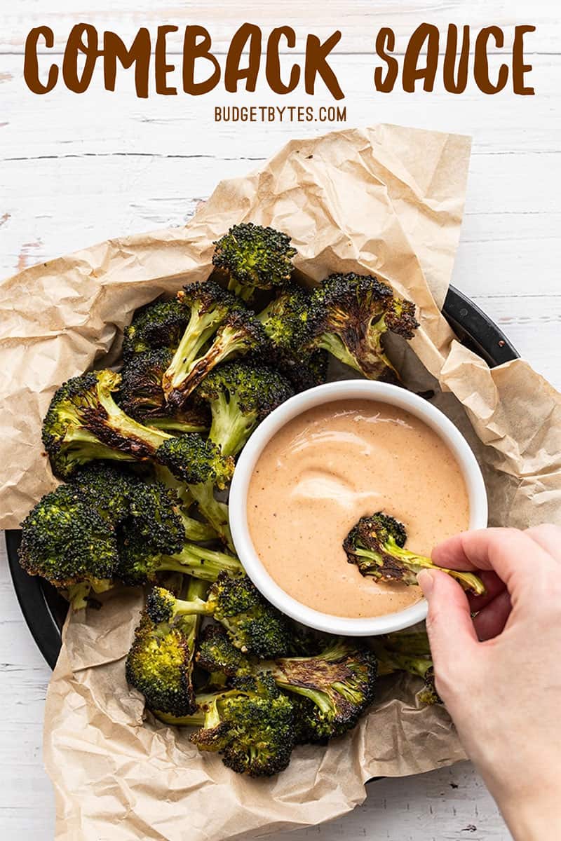 A hand dipping a piece of roasted broccoli being dipped into a bowl of comeback sauce