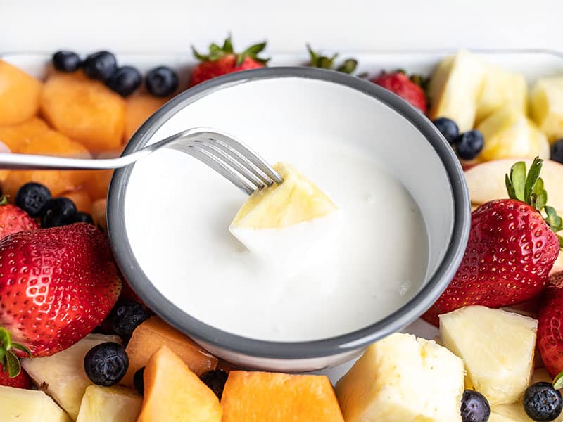 A fork dipping a piece of pineapple into the cottage cheese fruit dip, surrounded by fruit.