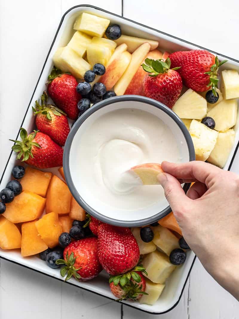 a hand dipping an apple slice into fruit dip on a tray of fruit