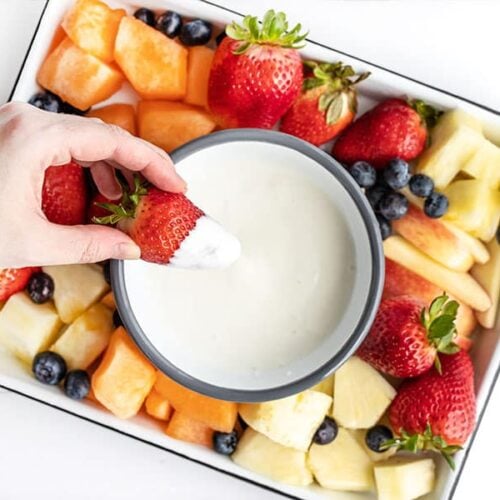 A hand dipping a fresh strawberry into the fruit dip on a fruit platter