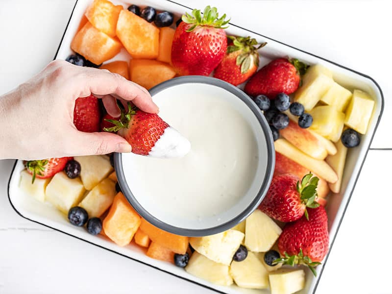 A hand dipping a fresh strawberry into the fruit dip on a fruit platter