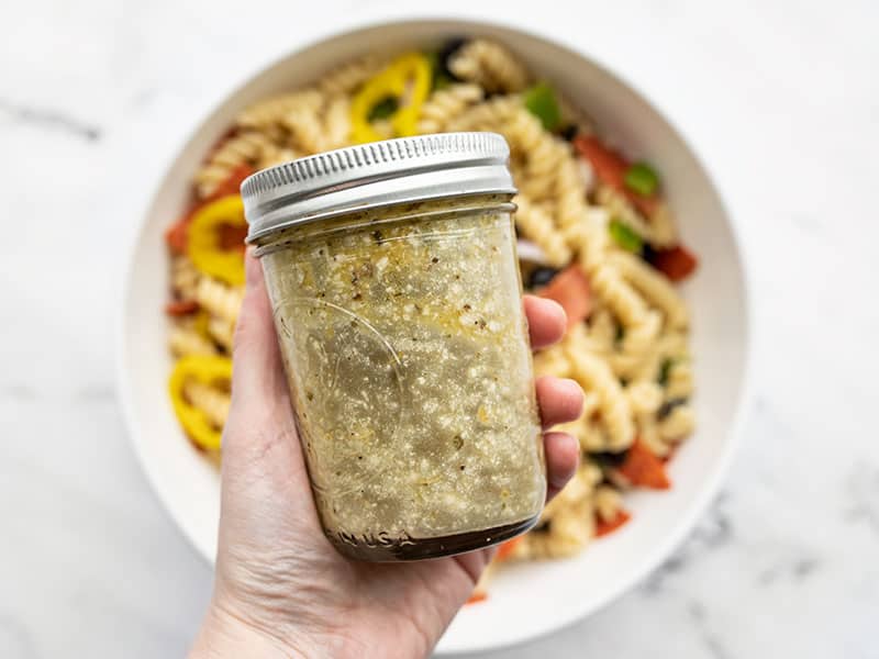 Shaken jar of dressing held over the bowl of pasta salad.