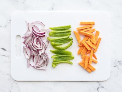 A white cutting board with sliced red onion, bell pepper, and sweet potato
