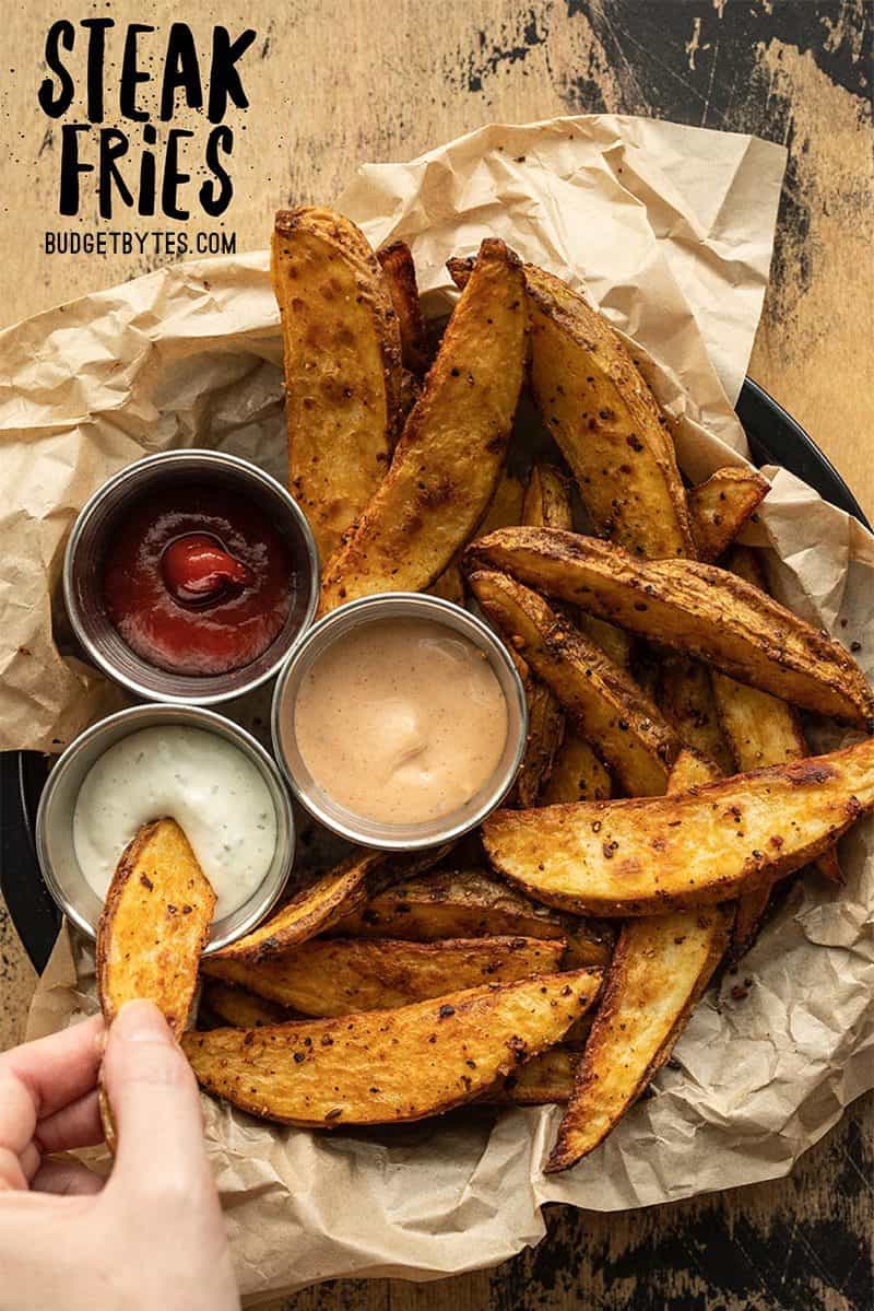 A hand dipping one steak fry from a plate of fries into a dish of sauce