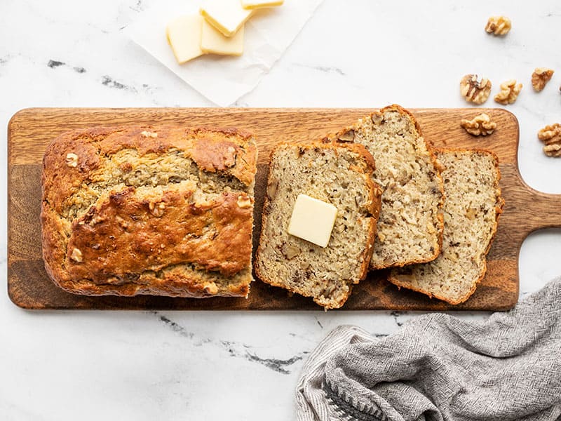 A half sliced loaf of yogurt banana bread on a wooden cutting board with one pat of butter in the center of one slice