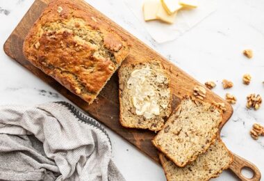 A loaf of yogurt banana bread on a wooden cutting board, a few slices scattered, one smeared with butter